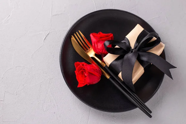 Romantic table setting. Black plate, knife, fork, box with present and red rose flower on textured grey background. Top view. Place fr text. Romantic postcard.