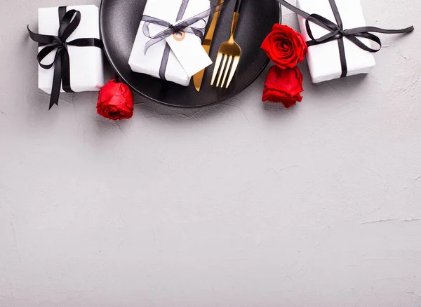 Romantic table  border. Black plate, knife, fork, boxes with presents and red roses flowers on textured grey background. Top view. Place fr text. Romantic postcard.