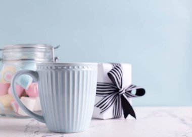 Romantik kartpostalı tebrik ederim. Mavi bardakta sıcak içecek, hediye kutusu ve mavi duvarda renkli marşmelovlarla dolu kavanoz. Seçici odaklanma. Metin için yer