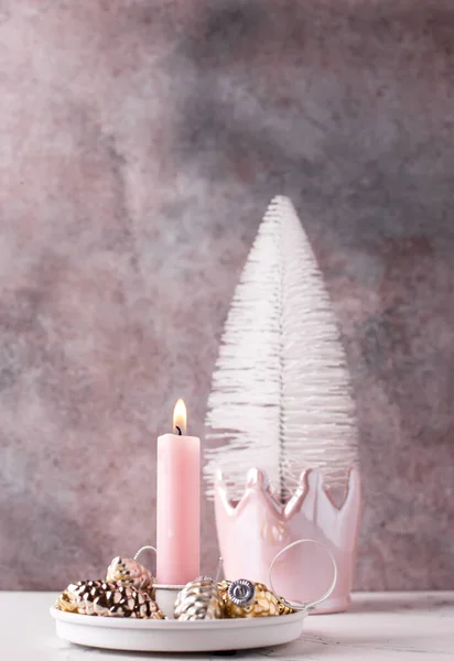 Pink burning candle and golden , silver decorative cones and white tree against pink textured wall. Selective focus is on candle. Place for text. Pink Christmas postcard.