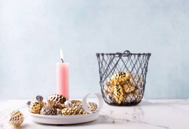 Kış tatili kartpostalı. Pembe yanan mum ve mavi desenli duvara karşı altın ve gümüş renkli dekoratif koniler. Seçici odaklanma. Metin için yer.