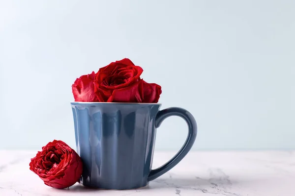 Romantic St. Valentine day postcard. Dark  blue cup with hot drink and  red roses flowers on white marble background against blue wall. Selective focus. Place for text.