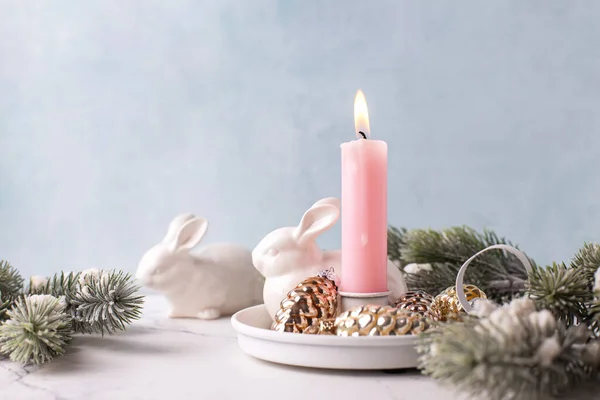 White bunnies, burning pink  candle , fir tree branches, decorative golden cones on white marble background against blue  textured wall. Rabbit is symbol of 2023.. Selective focus. Place for text.