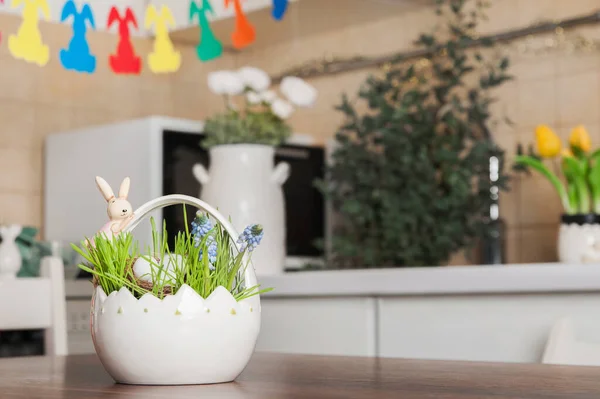 Basket with spring grass and Easter eggs on table on background of decorated kitchen. Spring kitchen decor for Easter.