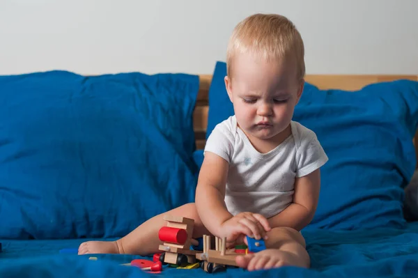 Toddler boy plays with wooden constructor train on bed close-up..