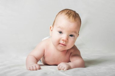 Nazik bir ifadeye sahip bir bebek dokulu bir battaniyenin üzerinde rahat bir şekilde uzanır, geniş gözlü bir merakla kameraya bakar. Yumuşak ışıklandırma rahatlatıcı atmosferi güçlendirir, bebekleri ön plana çıkarır.