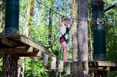 Açık hava macera parkına katılan genç bir kız, bir ip engelli parkurunda yol alıyor. Emniyet kemeri takıyor ve ipler arasında asılı ahşap platformu dikkatle geçiyor. Arka plan yemyeşil ile dolu, bu da ormanı işaret ediyor.