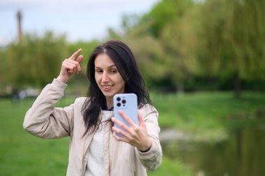 Gülümseyen kadın parkta video görüşmesi yapıyor ve yukarı doğru işaret ediyor..