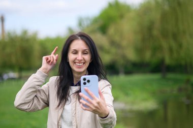 Gülümseyen kadın parkta video görüşmesi yapıyor ve yukarı doğru işaret ediyor..
