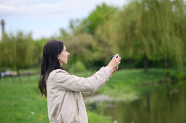Gülümseyen kadın parkta video görüşmesi yapıyor ve yukarı doğru işaret ediyor..