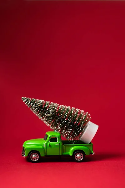 Green retro toy car carrying a Christmas tree on red background. Christmas and New Year celebration concept. Greeting card. Copy space, selective focus