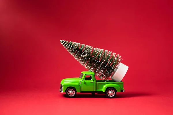 Green retro toy car carrying a Christmas tree on red background. Christmas and New Year celebration concept. Greeting card. Copy space, selective focus