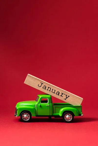 Green retro toy car carrying part of a wooden calendar with the month of January on red background. Christmas and New Year celebration concept. Greeting card. Copy space, selective focus