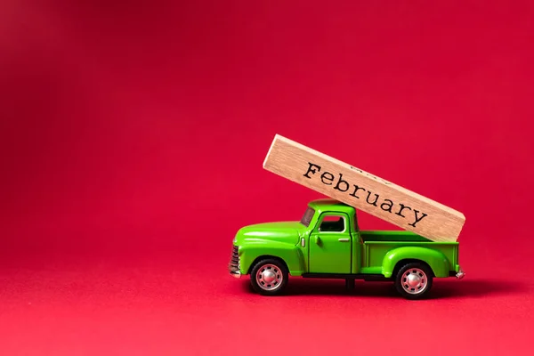 Green retro toy car carrying part of a wooden calendar with the month of February on red background. Christmas and New Year celebration concept. Greeting card. Copy space, selective focus