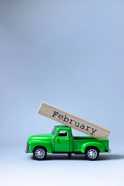 Green retro toy car carrying part of a wooden calendar with the month of February on grey background. Christmas and New Year celebration concept. Greeting card. Copy space, selective focus