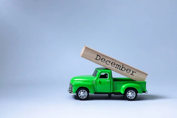 Green retro toy car carrying part of a wooden calendar with the month of December on grey background. Christmas and New Year celebration concept. Greeting card. Copy space, selective focus