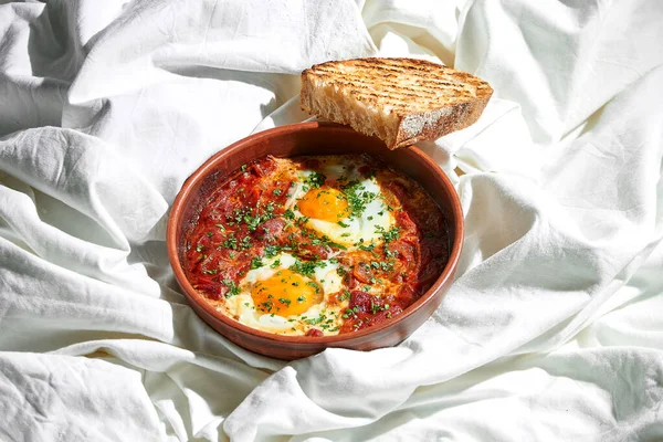 Breakfast in bed - scrambled eggs with Shakshouka tomatoes in a clay dish on a bed sheet
