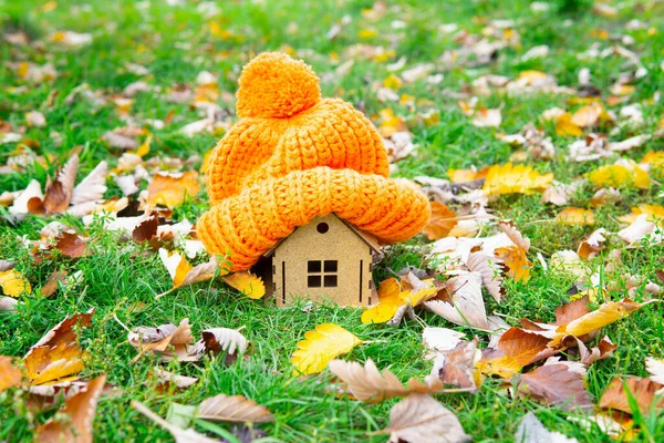 stock image Chunky knitted hat on a small toy house placed on a green lawn covered with autumn leaves. Staying warm at home in fall.