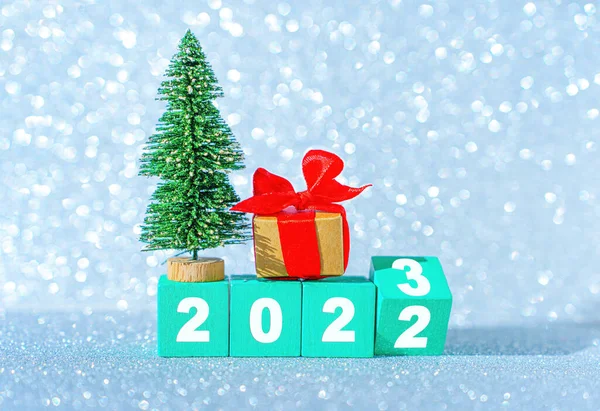 stock image New year 2023 beginning concept: half flipped wooden block, toy christmas tree and decorated gift box against a sparkling christmas background.