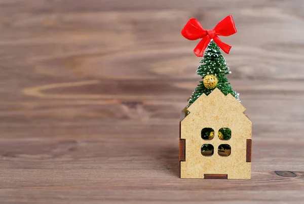 Christmas tree figurine with red bow on top placed inside a toy wooden house isolated on a wooden background. House decoration for the holidays season.