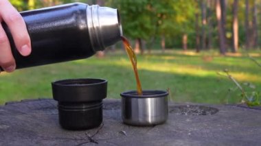 Man pours hot steaming coffee from the thermos into two cups placed on a large tree stump, puts the bottle next to it and closes it.