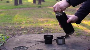Pair of thermos cups being placed on a large tree stump and filled with steaming coffee poured by a man from a thermos bottle.