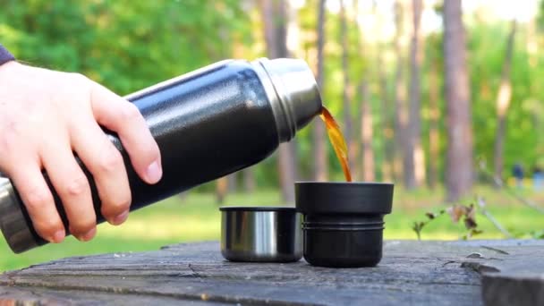 美しい秋の公園の大きな木の切り株に置かれた2つのカップに魔法瓶から熱いコーヒーを注ぐ男 ハイキングとコーヒーブレイクのコンセプト — ストック動画