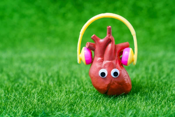 Music and nature concept: Heart-shaped character with googly eyes and headphones listening to music isolated on green grass background. Joy, relaxation, and carefree spirit design.