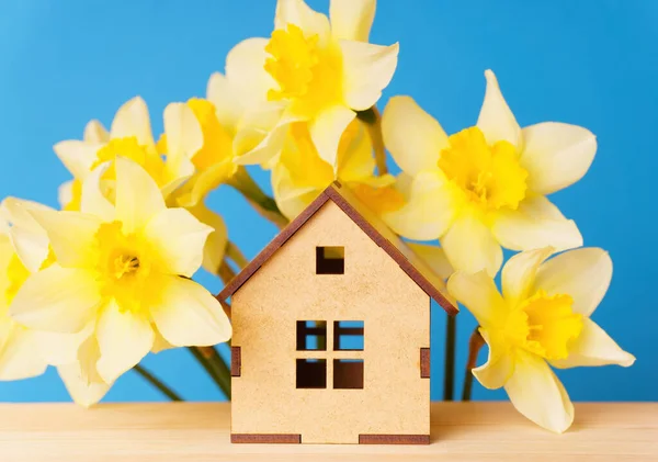 Miniature wooden house and a lovely bouquet of daffodils on a calming blue backdrop. Travel, retreats, and home decor related background.