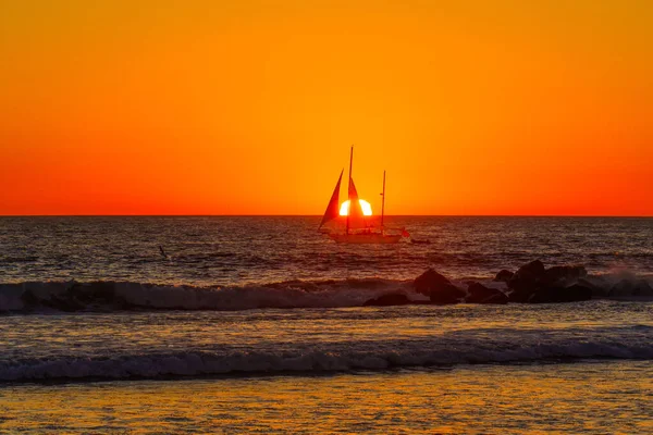 stock image Small sailboat sailing on the ocean waves against a setting sun standing out against the deep blue water, while the fiery oranges, yellows, and reds of the sunset sky create a stunning contrast.