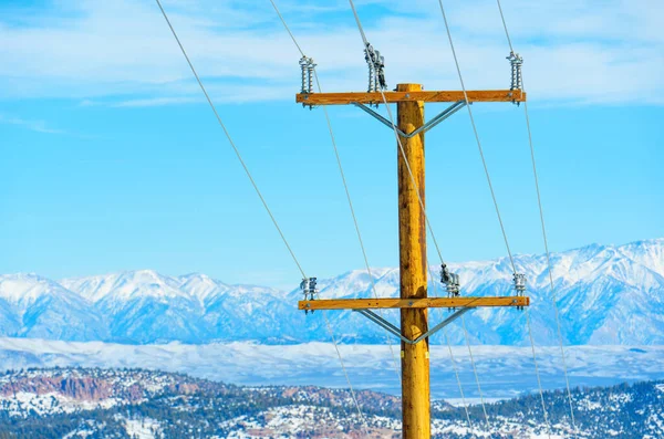Wooden power pole with wires against the backdrop of snow-covered mountain peaks and a clear blue sky. Energy, power supply, technology and engineering in mountainous regions.