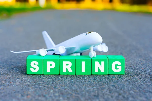Toy aircraft model placed on green wooden blocks spelling out the word \'Spring\', set against a rustic asphalt background leading to greenery. Creative spring season arrival concept.