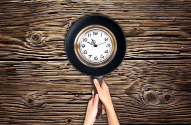 Yaşam, zaman ve günlük rutin kavramlar: Ellerin ahşap bir masa arka planına karşı kızarma yüzeyinde büyük bir vintage saat görüntüsü olan bir tavayı tutması.