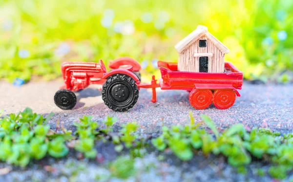 Small wooden house placed on a toy tractor trailer, parked on a paved road in the garden. Garden shed delivery concept.