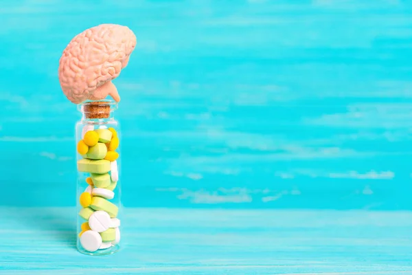 stock image Human brain model placed on top of a small glass bottle filled with colorful pills on a blue wooden background with copy space. Mental health and pharmaceuticals related concept.