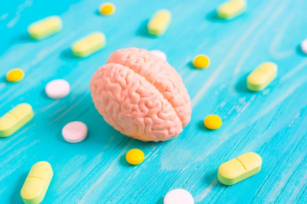Miniature human brain model alongside scattered yellow and white pills on a blue wooden table. Brain function, medication, treatment and preserving concept.