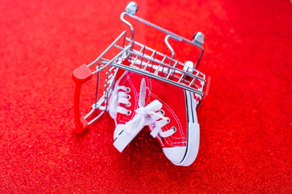 Pair of tiny red canvas shoes placed inside an overturned miniature push trolley, set against a glittering red backdrop. Gift-giving, holidays, e-commerce or fashion marketing backdrop.