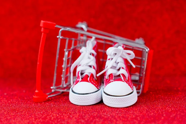 Pair of toy-size red canvas shoes resting inside an overturned miniature shopping cart, set against a glittering red background. Shopping, gift giving, holidays, and e-commerce related concept.