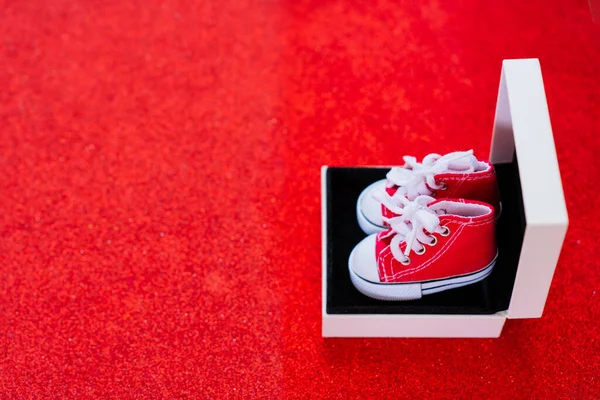 Pair of toy-size red canvas shoes resting inside a small gift box, set against a glittering red background. Shopping, gift giving, holidays, and e-commerce related concept.