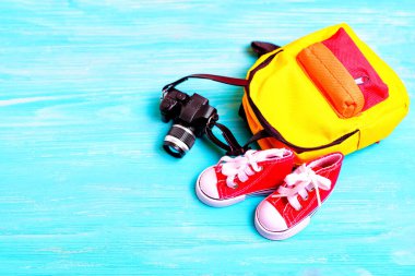 Bir çift küçük kırmızı tuval ayakkabı, renkli sırt çantası ve mavi ahşap arka planda fotokopi alanı olan bir oyuncak kamera. Yaratıcı okula dönüş veya seyahatle ilgili projeler.