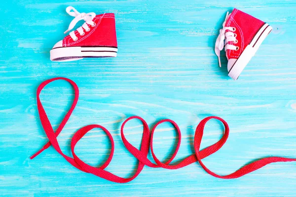 Little red canvas sneakers placed next to the word LOVE made of red laces on a blue wooden background.