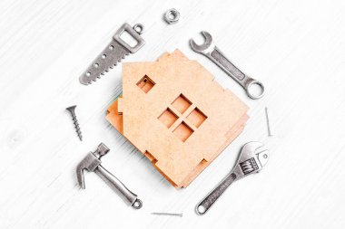 Parçalanmış ahşap ev modelinin üst görüntüsü ve çekiç, testere, anahtarlar, fındık ve çivi gibi çeşitli el aletlerinin küçük çelik kopyaları beyaz ahşap bir arka planda düzenlenmiştir..