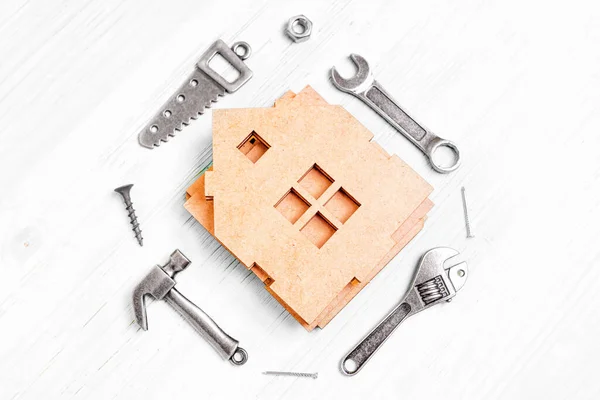 stock image Top view of a disassembled wooden miniature house model and small steel replicas of various hand tools such as a hammer, saw, wrenches, nuts and nails, arranged on a white wooden background.