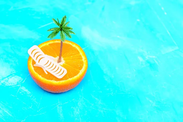 Halved orange fruit with a miniature lounge chair and a toy palm tree placed on a beautiful turquoise background with copy space. Tropical summer vacation and travel promotions concept.