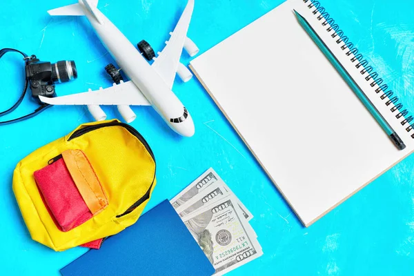 Creative flat-lay related to flight preparation: Toy plane model, notebook with a pencil, vibrant backpack, passport with cash and tiny camera arranged on a blue background.