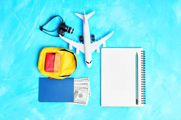 The concept of getting ready for a plane trip: Toy plane model, notebook with a pencil, vibrant miniature backpack, passport with cash and tiny camera arranged on a blue background.