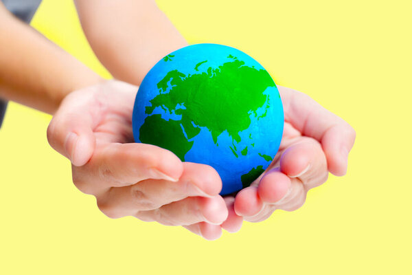 Крупный план женских рук, мягко держащих голубую и зеленую сферическую модель Земли на желтом фоне.