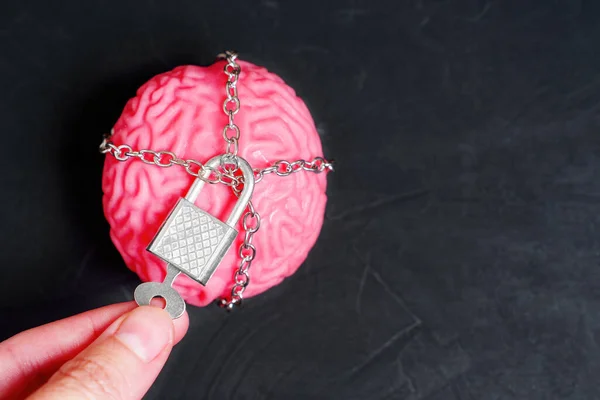 Human brain wrapped in chains and secured with a padlock. Hand uses a key on the padlock.