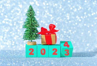 2023 'ten 2024' e geçiş: yarım ters çevrilmiş ahşap blok, oyuncak Noel ağacı ve parlak bir arka planda süslemeli hediye kutusu.