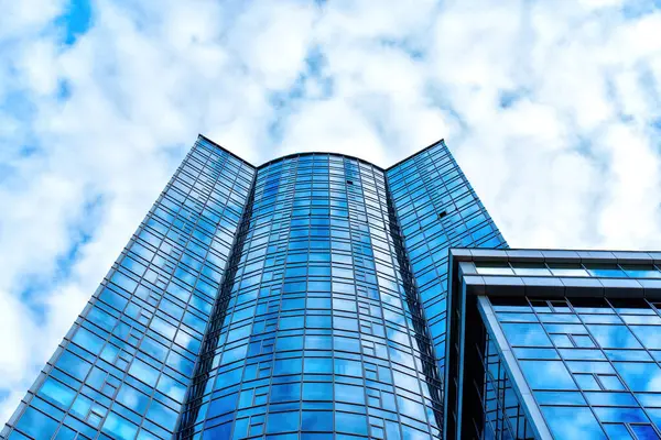 Wide angle view from below of a sleek glass office building set against a backdrop of clear blue sky adorned with fluffy white clouds.